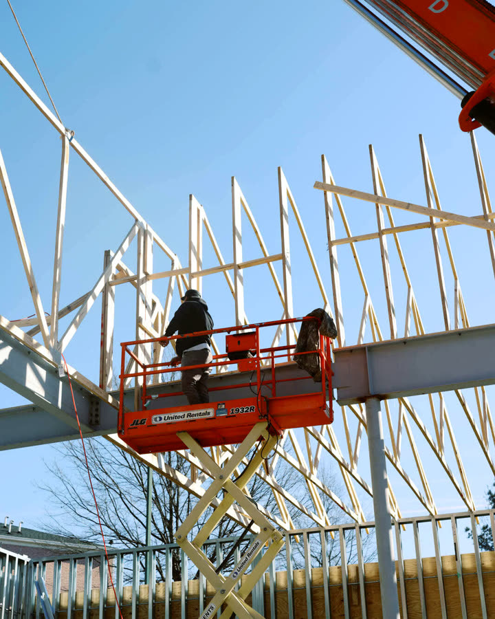 A worker on a scissor lift setting roof trusses on a steel frame
